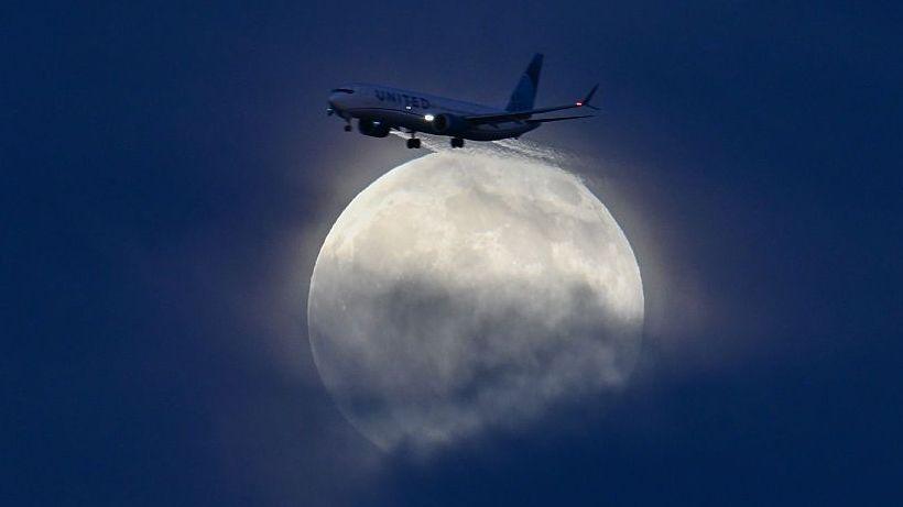A large, glowing white moon is rising through blue cloud as a plane flies. The plane is at the top of the moon and the sky is a dark blue behind it.